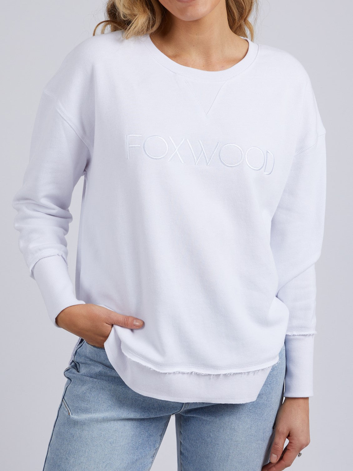 A woman wears the Foxwood Simplified Crew, a relaxed fit white 100% cotton sweatshirt with "FOXWOOD" embroidered on the front, paired with light blue jeans and posing with one hand in her pocket against a plain background.