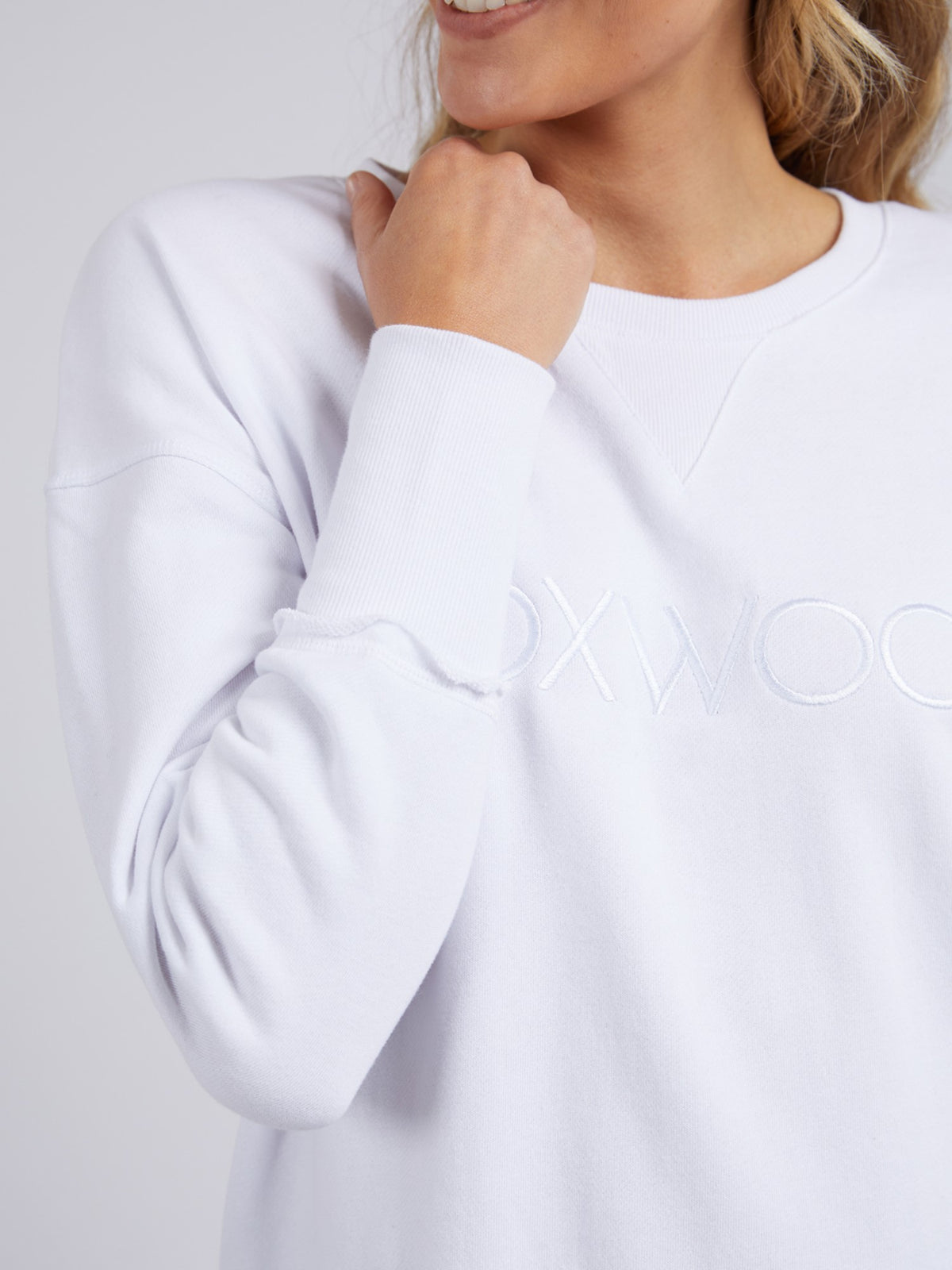 A woman smiles in the Foxwood Simplified Crew, a white sweatshirt with an embroidered logo. The image focuses on her relaxed pose, highlighting the comfortable fit and stylish details of this casual top.