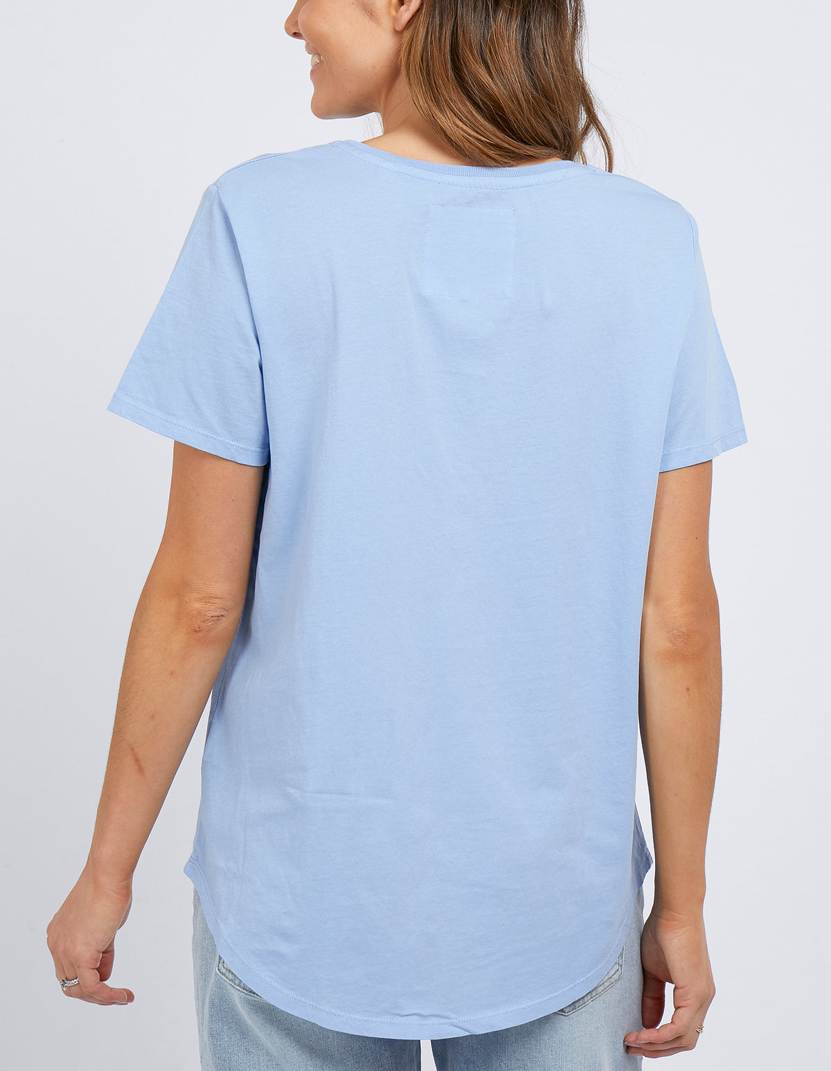 A woman with light brown hair is shown from the back, smiling and slightly raising her right hand. She wears a light blue 100% cotton Foxwood Tee—a casual staple—and light jeans.