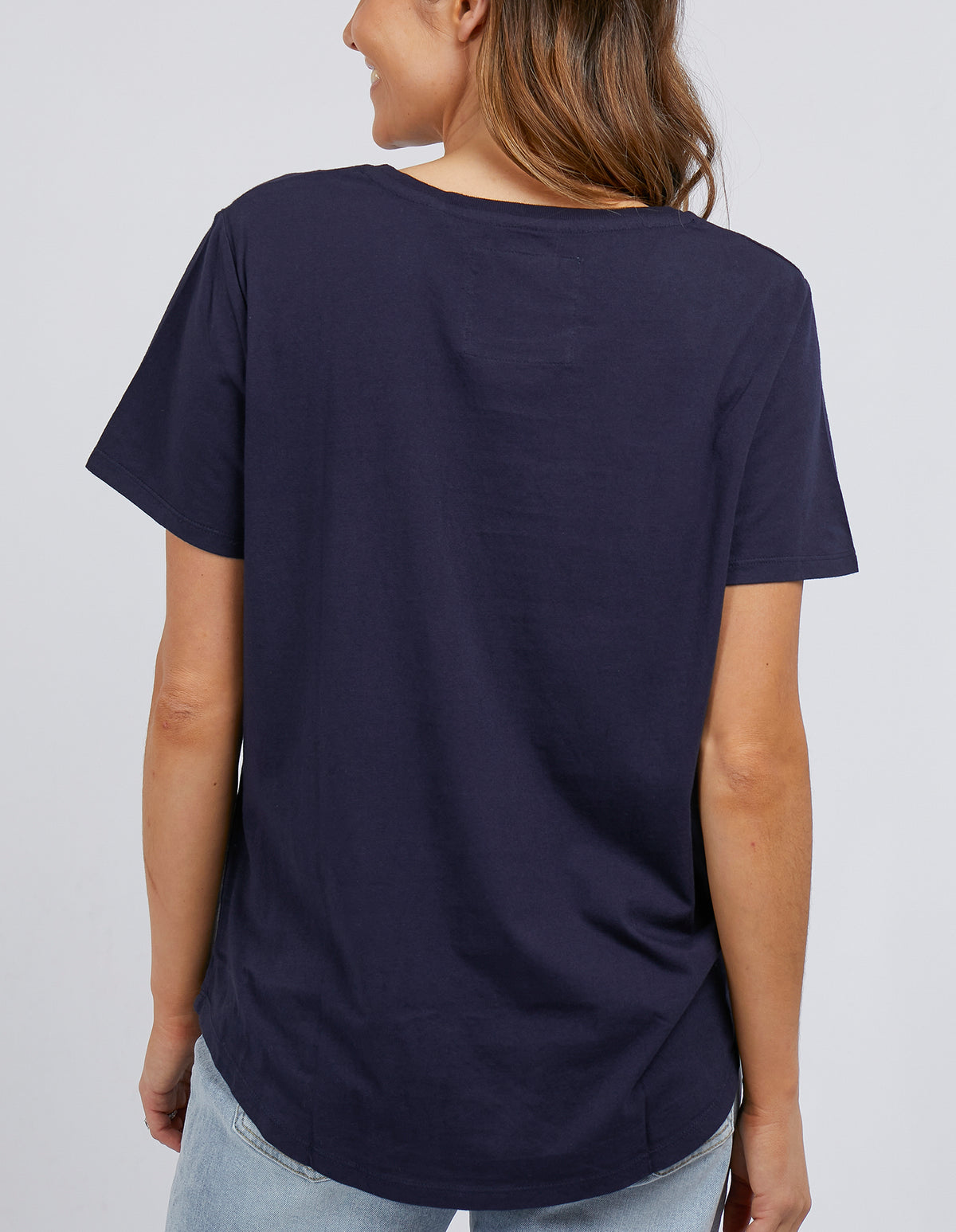 A person with long hair wears a Foxwood Tee, a navy 100% cotton shirt, paired with light blue jeans. With their back to the camera against a plain light gray background, they embody effortless style.
