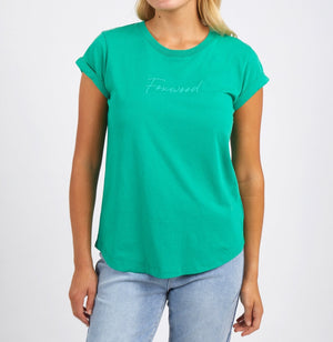 A woman models the Foxwood Signature Tee, a bright green 100% cotton casual top with "Foxwood" in cursive on the chest. She pairs it with light blue jeans and stands against a plain white background.