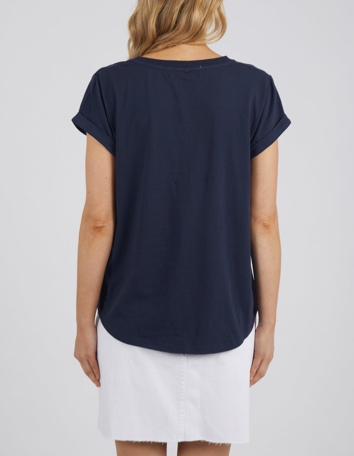 A person with long hair is seen from behind wearing a Foxwood Manly Vee Tee in dark blue with rolled cuff sleeves, paired with a white skirt. They're framed against a plain gray background creating a contemporary look.