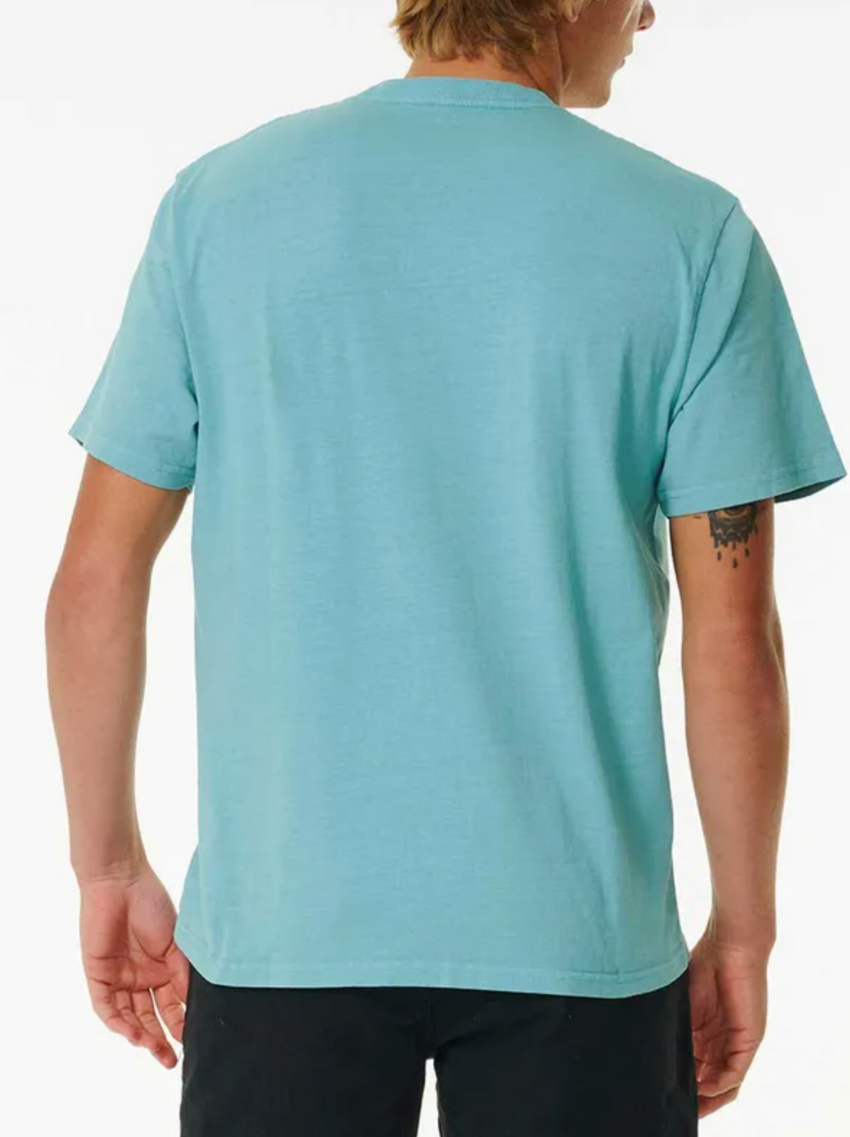 A person with light hair, facing away from the camera, sports a light blue Rip Curl Plain Wash Tee in a relaxed fit alongside black pants. A small tattoo is visible on their left arm, complementing their laid-back style.