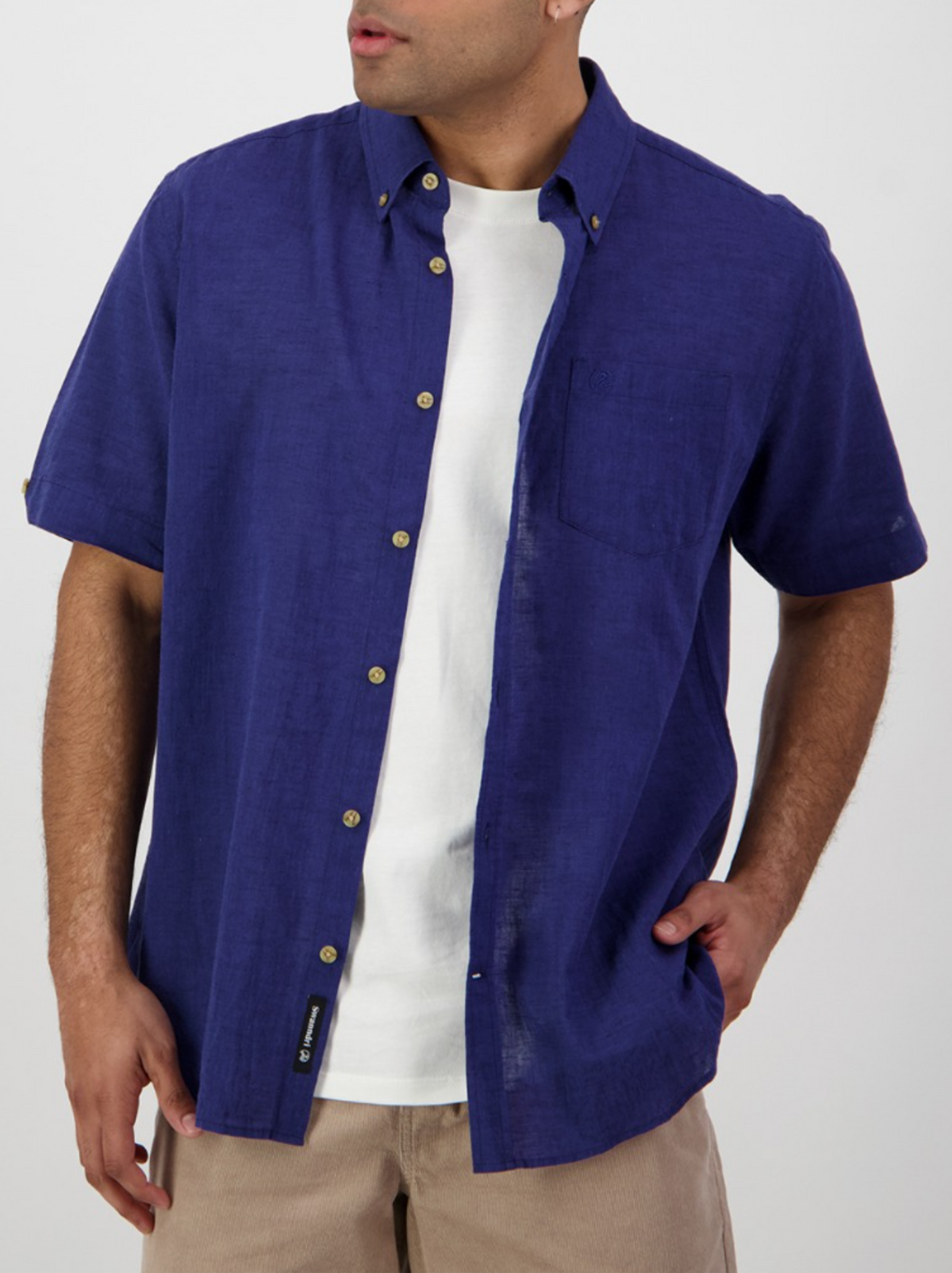 A man wears the Swanndri Lowell v2 Short Sleeve Shirt in bright blue over a white T-shirt and beige pants, standing with one hand in his pocket against a plain background.