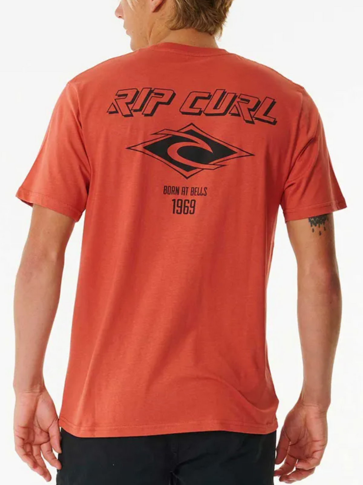 A person is seen from behind wearing the Rip Curl Fade Out Icon Tee, an orange 100% cotton shirt featuring a black Rip Curl logo, diamond-shaped graphic, and vintage “BORN AT BELLS 1969” text on the back.