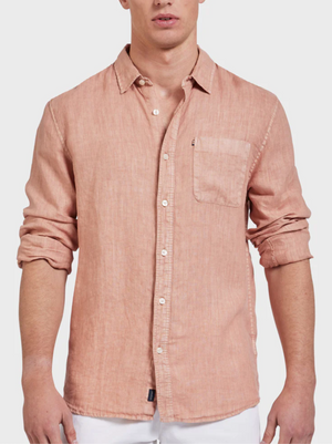 A man wears the Academy Brand Hampton Linen Shirt in light pink with white buttons and a chest pocket, paired with white pants. The shirt’s sleeves are rolled up for a relaxed vibe.
