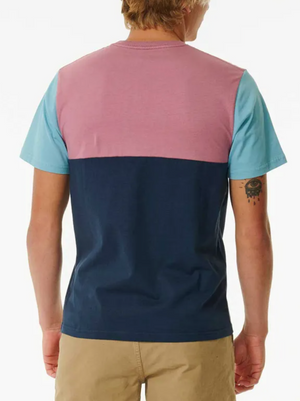 A person with short light brown hair and a left arm tattoo is seen from behind wearing the Rip Curl Surf Revival Custom Tee—100% cotton, featuring a pink upper back, light blue sleeves, navy lower half—and tan shorts.