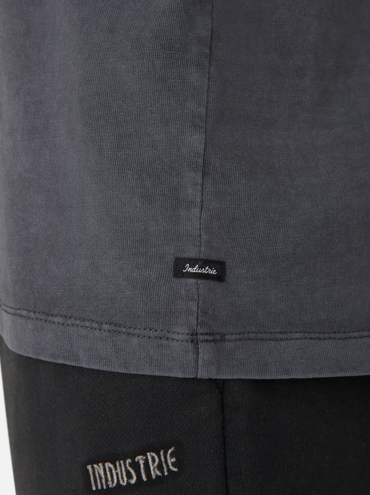 Close-up of a person's side wearing the oversized Industrie The Del Sur Tee in dark gray, with a small black label near the hem. This 100% cotton T-shirt effortlessly pairs with black pants featuring a white-stitched "Industrie" logo near the waistband.