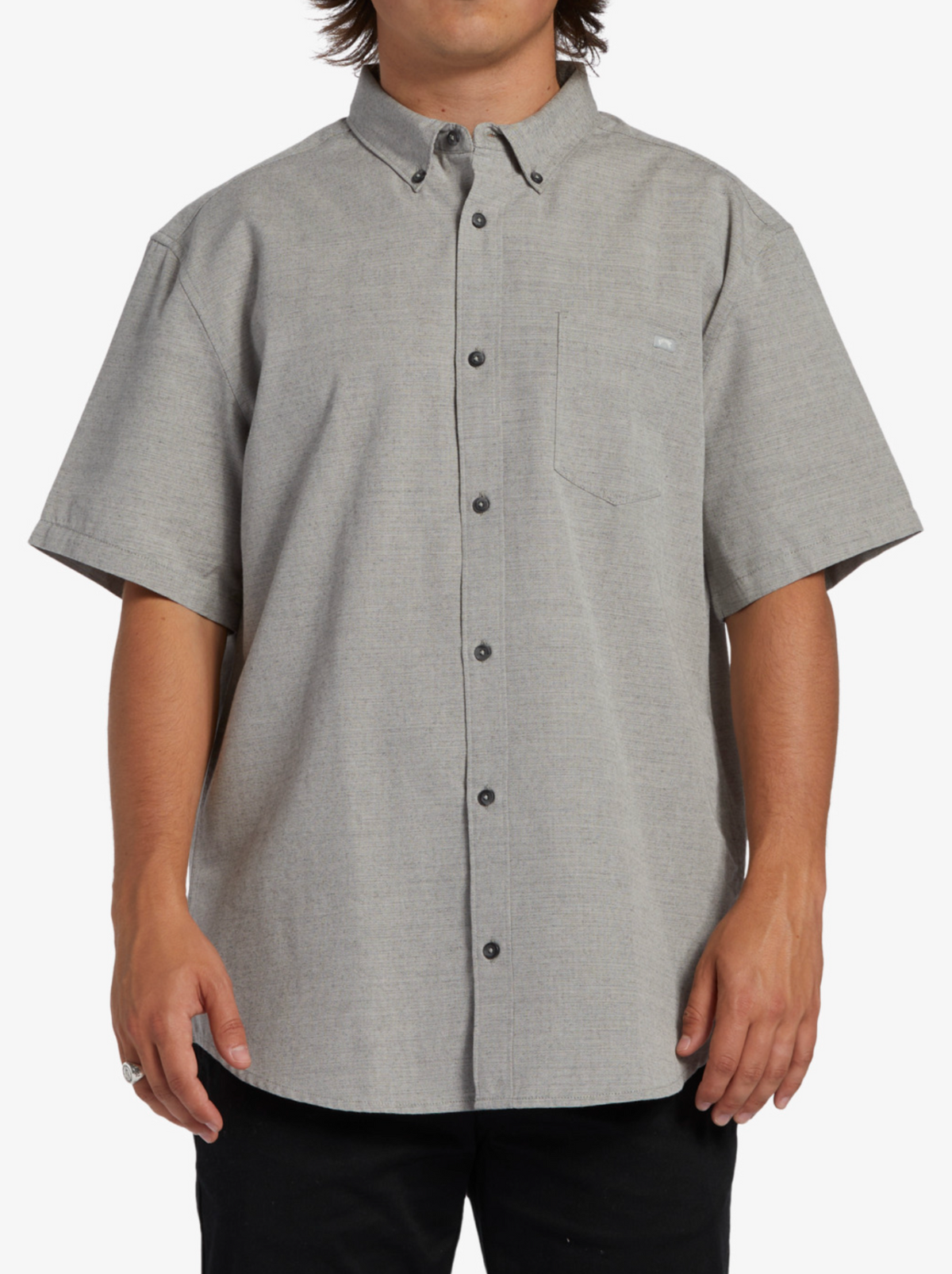 A person wears the Billabong All Day Short Sleeve Shirt in light gray, made from cotton polyester woven fabric, with a collar and chest pocket. Its subtle pattern pairs well with black pants, all on a plain white background.