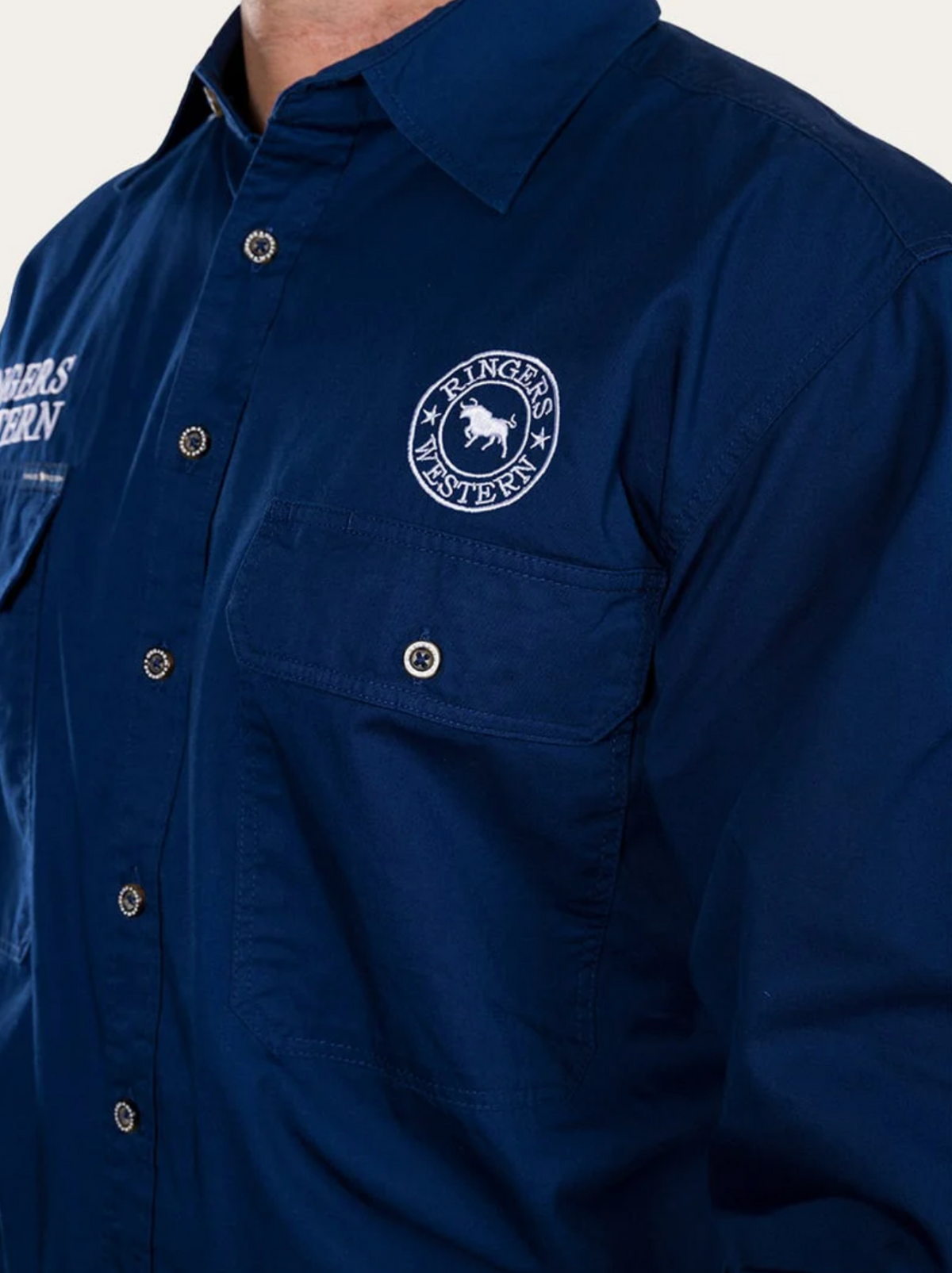 A person wearing a dark blue Ringers Western Hawkeye Embroidered Shirt with a "RINGERS WESTERN" logo and horse emblem on the chest pocket, white stitching, visible buttons, and UPF50+ protection.