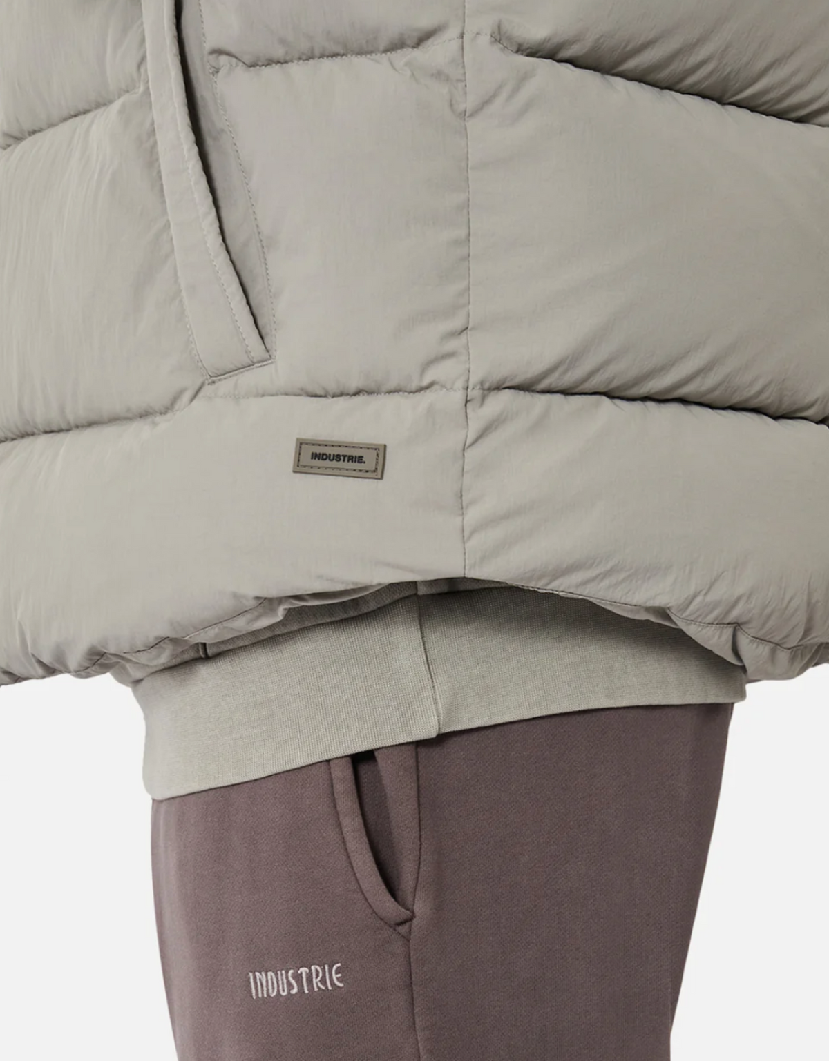 Close-up of a person wearing a light gray puffer vest with thick quilted padding and mauve sweatpants. The vest is zipped, featuring a small "INDUSTRIE" tag, and the sweatpants have "INDUSTRIE" written on the left side near the pocket. The background is plain white.