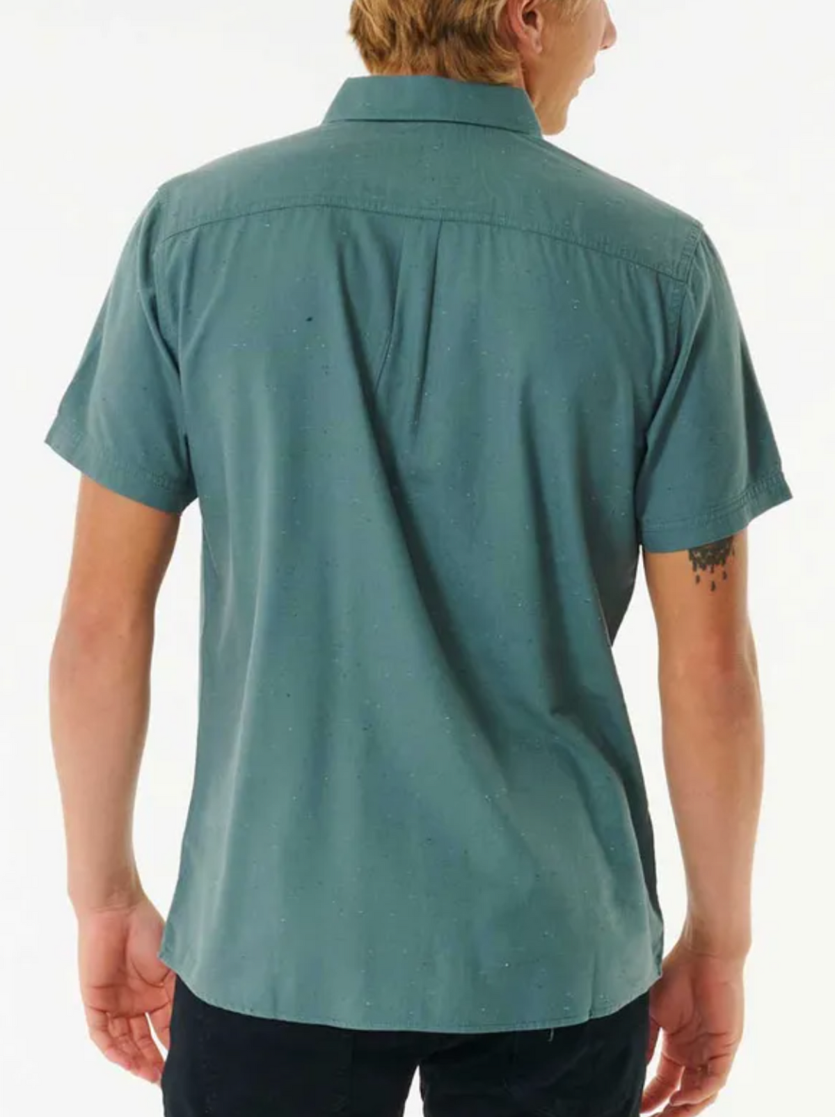 A man, with short light brown hair and a tattoo on his upper right arm, stands facing away in dark pants and a Rip Curl Ourtime Short Sleeve Shirt in teal.