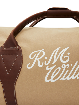 Close-up of the RM Williams Sorrento Ute Bag, showcasing its beige water-repellant utility canvas body with brown leather handles and trim, accented by white decorative stitching. The bag features "R.M. Williams" printed in white cursive lettering.