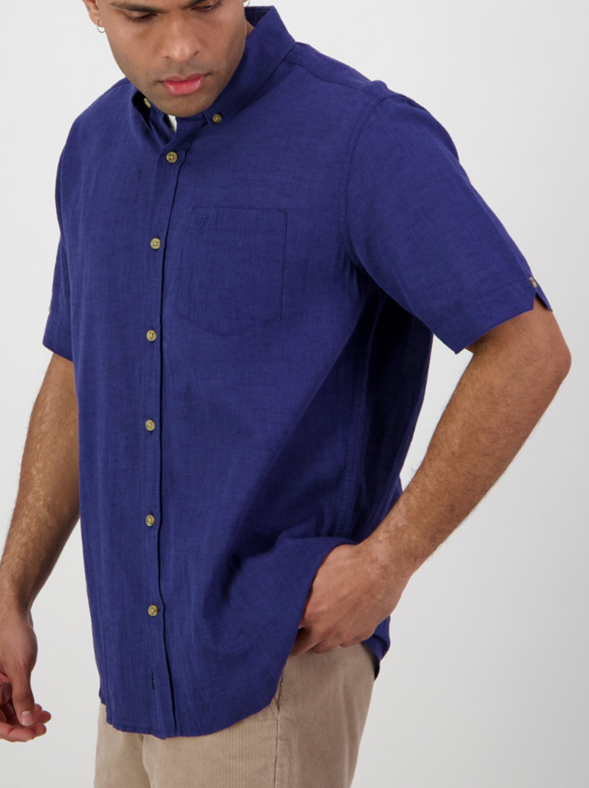 A man wears the Swanndri Lowell v2 Short Sleeve Shirt, a dark blue button-up with a button-down collar, and beige pants. He looks down with one hand in his pocket against a plain light background.