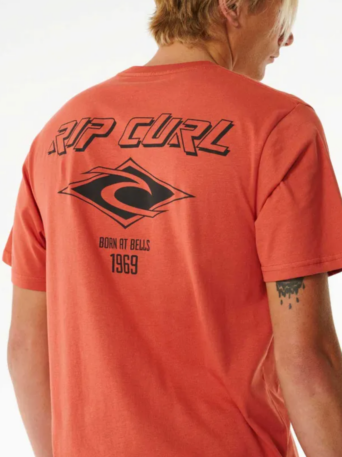 Wearing a 100% cotton orange Rip Curl Fade Out Icon Tee, featuring a large black logo on the front and "RIP CURL" with "BORN AT BELLS 1969" on the back. Part of a geometric tattoo is visible on the wearer's arm.
