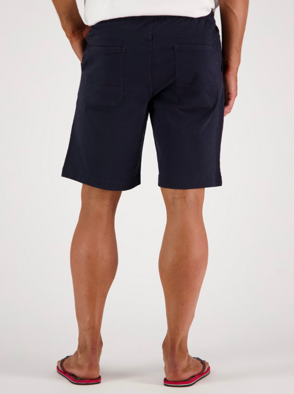 A person is seen from behind wearing a white shirt and Swanndri Clevedon Stretch Canvas Shorts in navy blue, featuring an elastic waist and back pockets. Red flip-flops complete the relaxed ensemble as they face away on a light background.