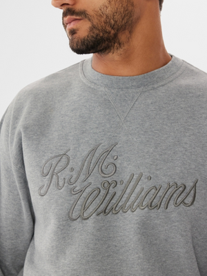 A person with short dark hair and facial hair stands against a plain backdrop wearing the RM Williams R.M.W Script Crew Neck, a gray jumper stylishly embroidered on the front, enhancing their casual yet classic look.