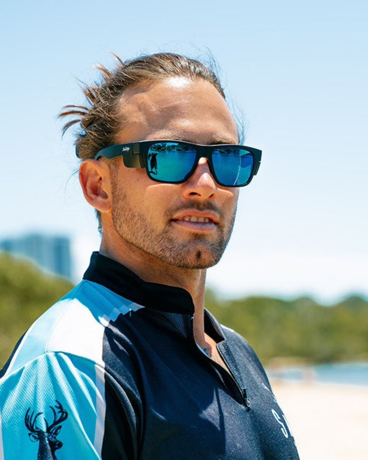A man with tied-back hair and a short beard wears SafeStyle Fusions Matte Black Frame Mirror Blue Polarised Lens sunglasses and a blue sports shirt with a deer head logo, standing outdoors on a sunny day with trees and buildings in the background.