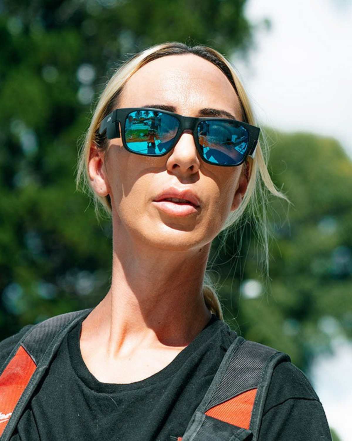 A light-haired person in a ponytail, wearing SafeStyle Fusions Matte Black Frame sunglasses with Mirror Blue Polarised Lens and a black shirt, stands outdoors among green trees and blue sky, carrying a backpack.