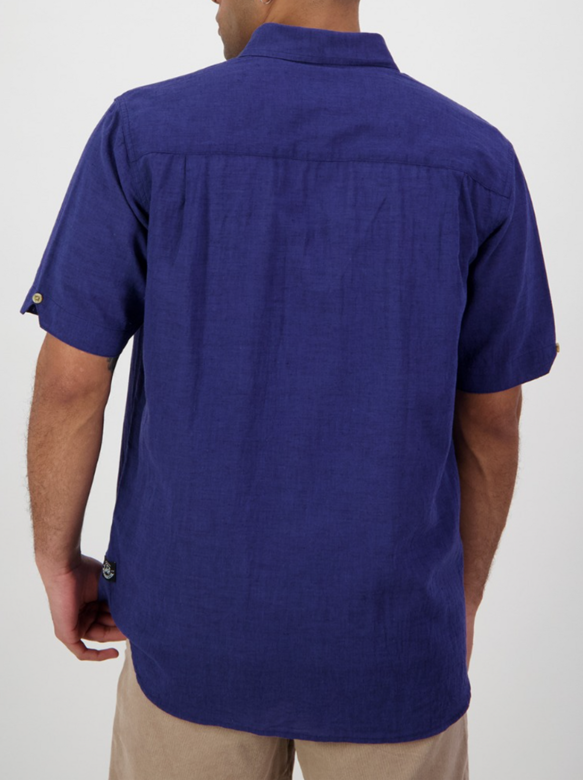 A person is shown from the back wearing a Swanndri Lowell v2 Short Sleeve Shirt in deep blue, featuring a button-down collar, button tabs on the sleeves, lightweight linen fabric, and paired with light-colored pants against a plain background.