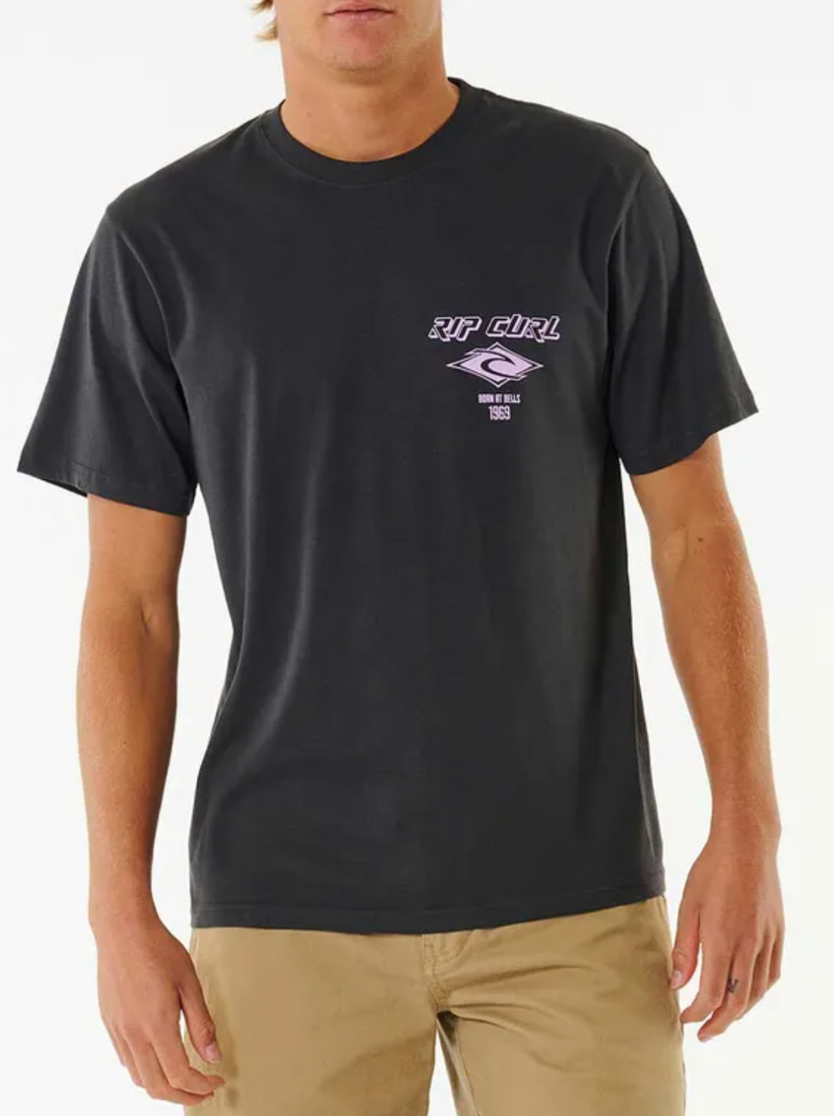 A person in a black Rip Curl Fade Out Icon Tee, featuring a small chest logo and vintage style, paired with khaki pants. Made of 100% cotton, they stand out against the white backdrop.