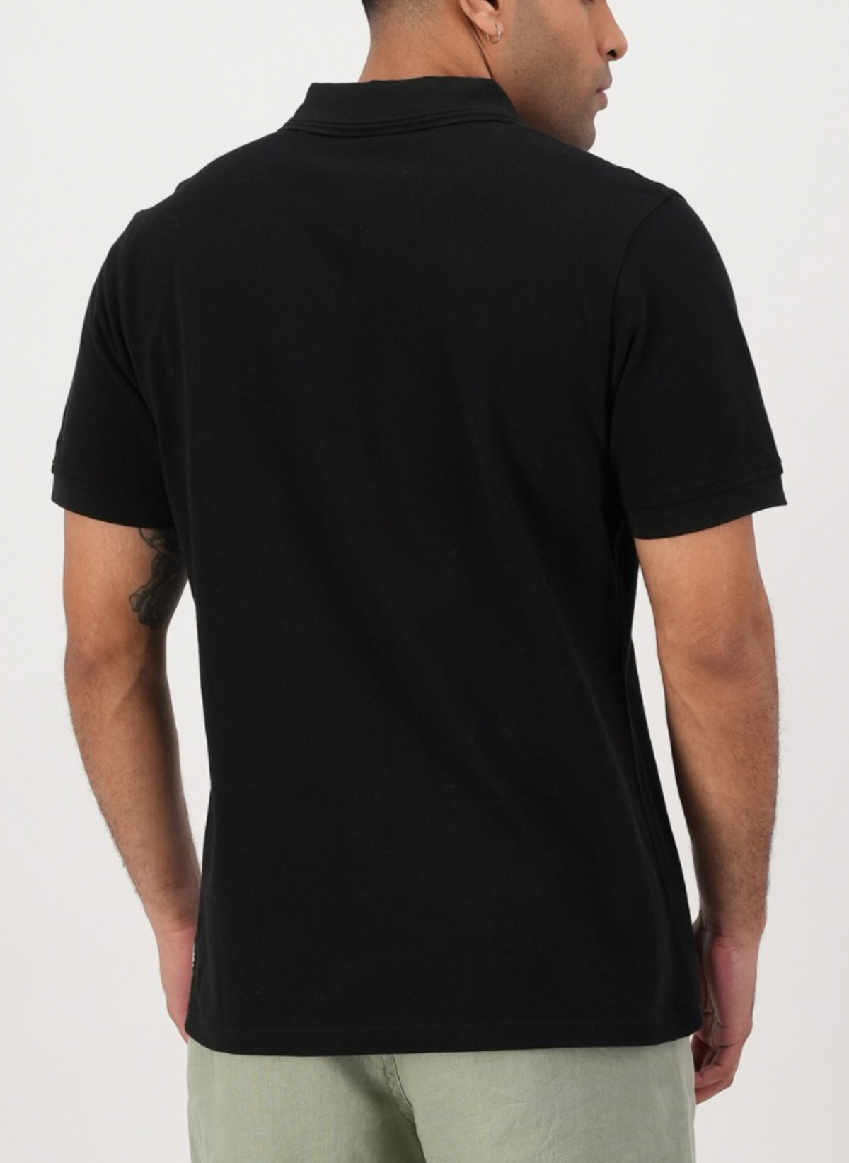 A person is wearing the black Swanndri Highgrove Polo, crafted from luxurious cotton pique, and light-colored pants. They stand against a plain white background with their back to the camera and arms relaxed by their sides.