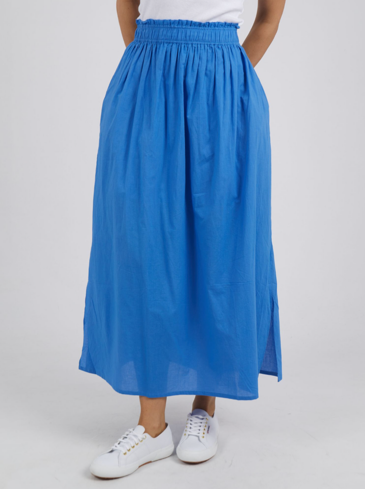A person is wearing a bright blue, ankle-length Foxwood Charli Skirt with an elastic waistband, paired with a white top. The skirt, made from 100% cotton, has a slightly flared design and a side slit. The person is also wearing white sneakers. The background is plain white.