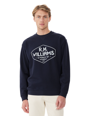 A person with light brown hair stands against a white background, wearing light-colored pants and the RM Williams Gladstone Crew Neck—a relaxed-fit dark blue sweater featuring a white R.M.Williams graphic that reads "5 Percy St Prospect S.A.