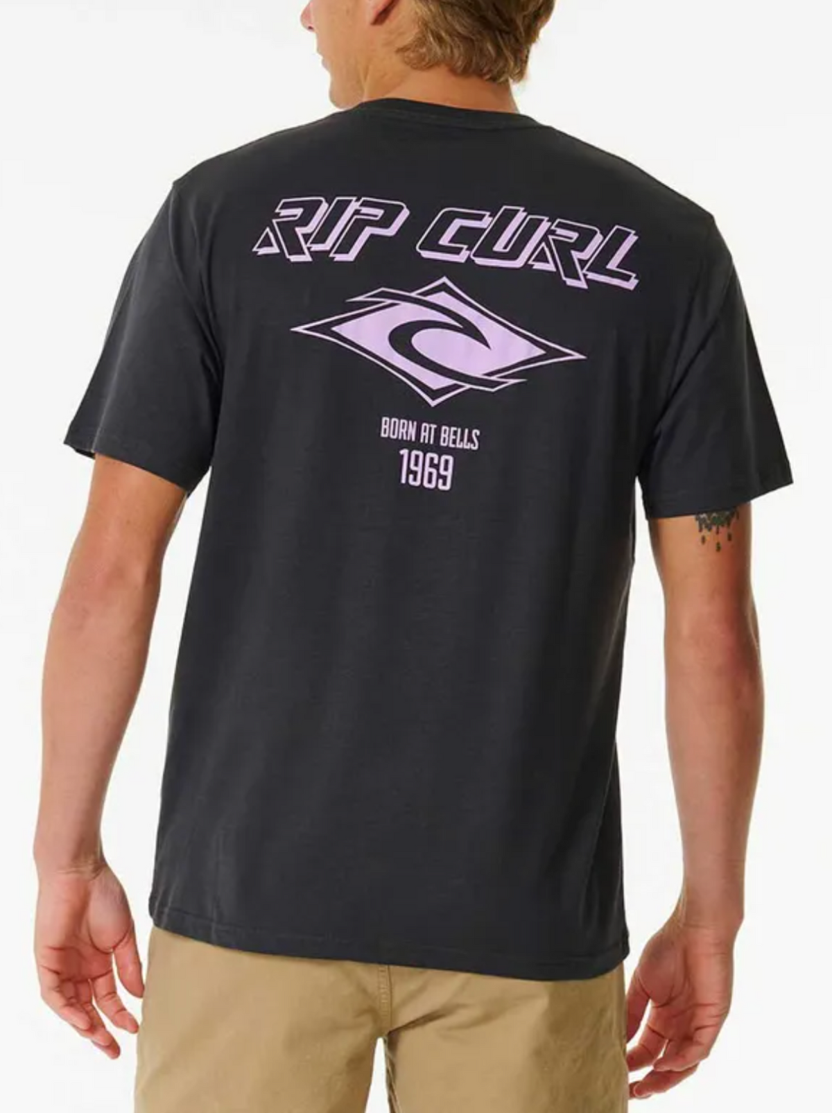 Wearing the black Rip Curl Fade Out Icon Tee with light pink "Rip Curl" text and "Born at Bells 1969" on the back, exudes a throwback style. The iconic logo complements their light brown shorts, making this 100% cotton tee an ideal vintage-inspired outfit choice.