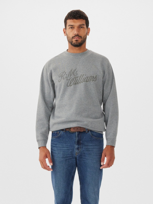 A person with short dark hair and a beard wears the RM Williams R.M.W Script Crew Neck in gray, showcasing "R.M. Williams" embroidery. They pair it with blue jeans and a brown belt, standing against a plain white background to highlight the stylish cotton blend fabric.