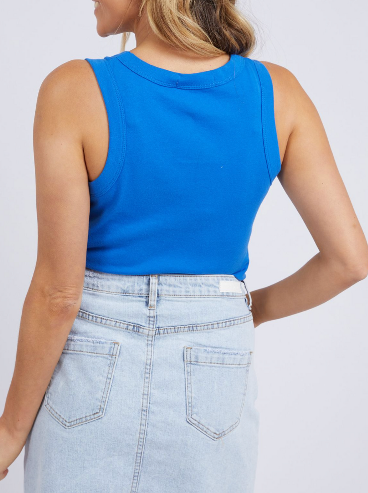 A woman, seen from the back with her head turned, wears the bright blue Foxwood Esme Tank styled with a light-wash denim skirt.