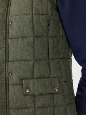 Close-up of a person wearing the RM Williams Wilpena Creek Vest, a quilted green piece crafted from recycled polyester with pocket and button details, over a navy long-sleeve shirt. The sporty vest is partially zipped, highlighting its textured fabric.