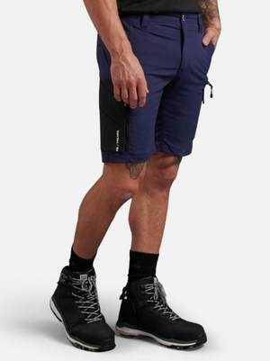 Wearing King Gee Trademark Cargo Short in navy blue stretch ripstop with a black side pocket and belt, they pair it with black socks and hiking boots featuring white soles. Their tattooed left arm stands out against the white background, highlighting their adventurous style.