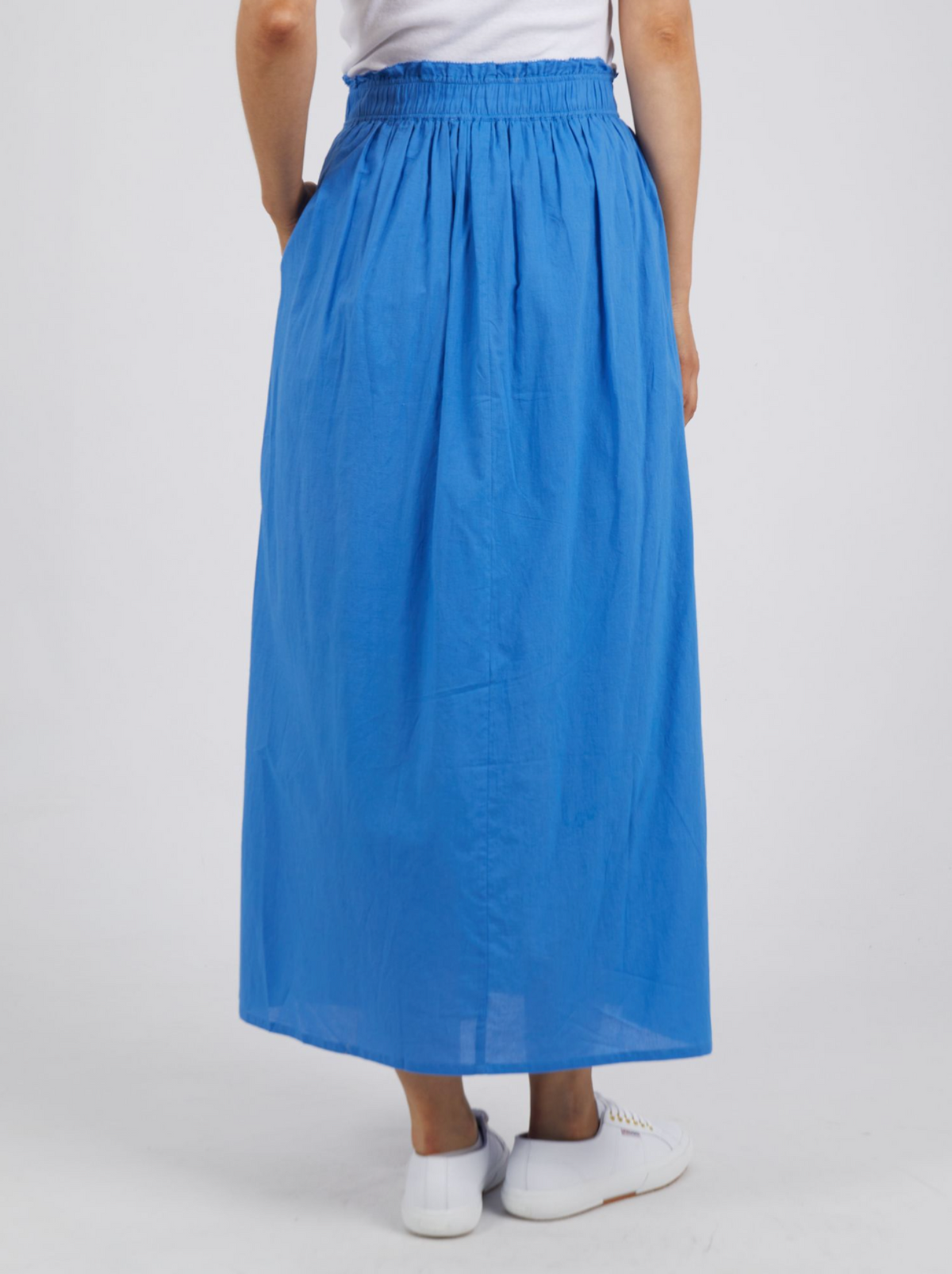 A person is shown from the back, wearing a blue Foxwood Charli Skirt made of 100% cotton with an elastic waistband. The individual is also wearing a white top and white sneakers, standing against a plain, light background.