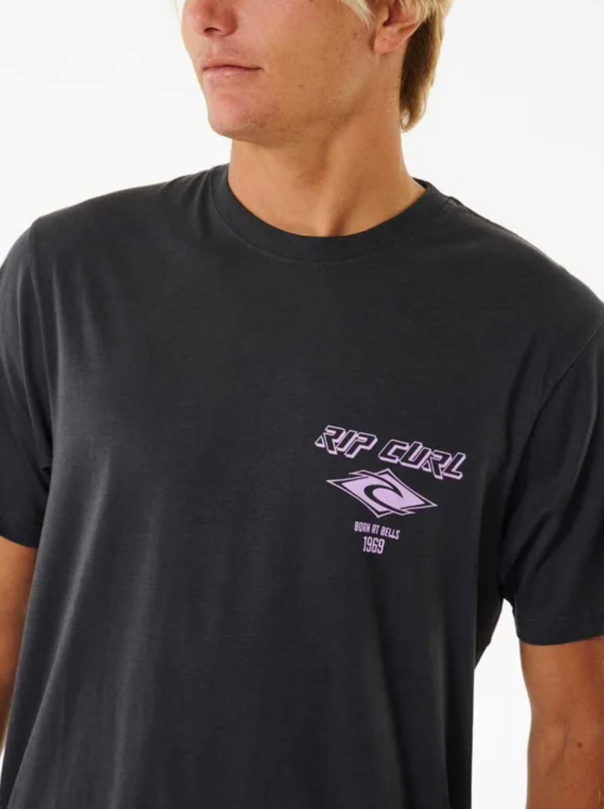 The person is wearing the Rip Curl Fade Out Icon Tee, a retro black T-shirt with a purple "Rip Curl" logo and "Born at Bells 1969" on the left chest, made from 100% cotton. It pops against a plain white background.