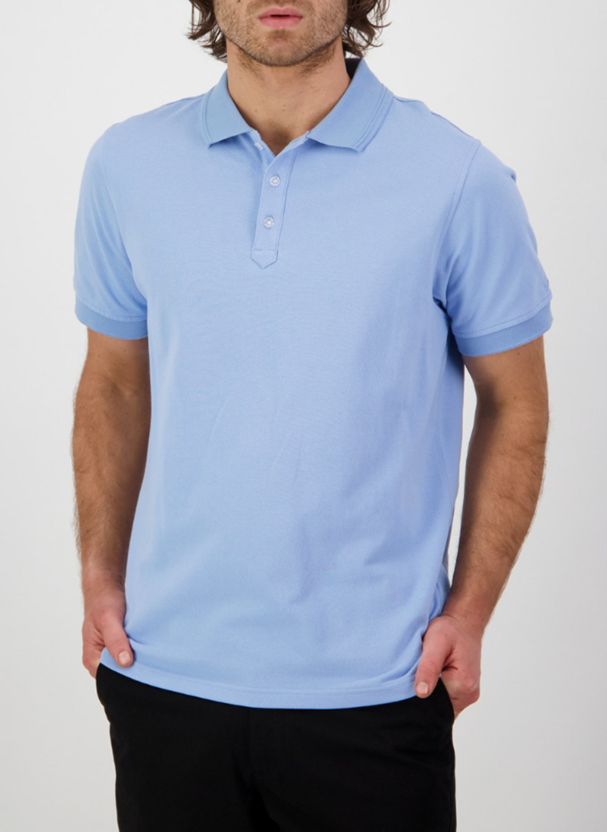 A man in a Swanndri Highgrove light blue polo shirt, crafted from combed cotton pique, stands confidently against a white background with his hands casually in his pockets.