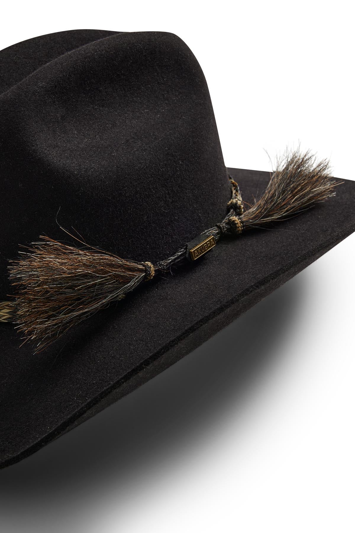 A close-up of the Akubra Rough Rider hat in black felt, featuring a Cattleman crown, decorative braided band with brown and black tassels, a metallic tag, all set against a white background.