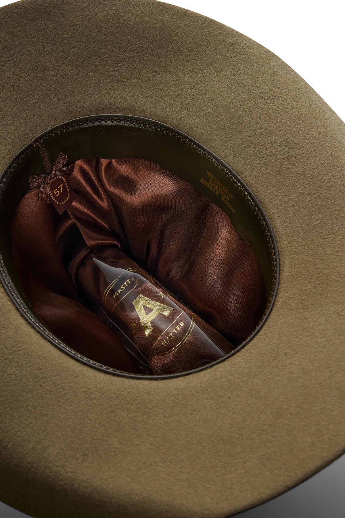 A brown glass bottle labeled "A" sits inside the satin-lined interior of an olive-green Akubra Rough Rider hat, with a gold size tag marked "57" attached to the inner band.
