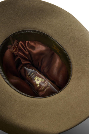 A brown glass bottle labeled "A" sits inside the satin-lined interior of an olive-green Akubra Rough Rider hat, with a gold size tag marked "57" attached to the inner band.