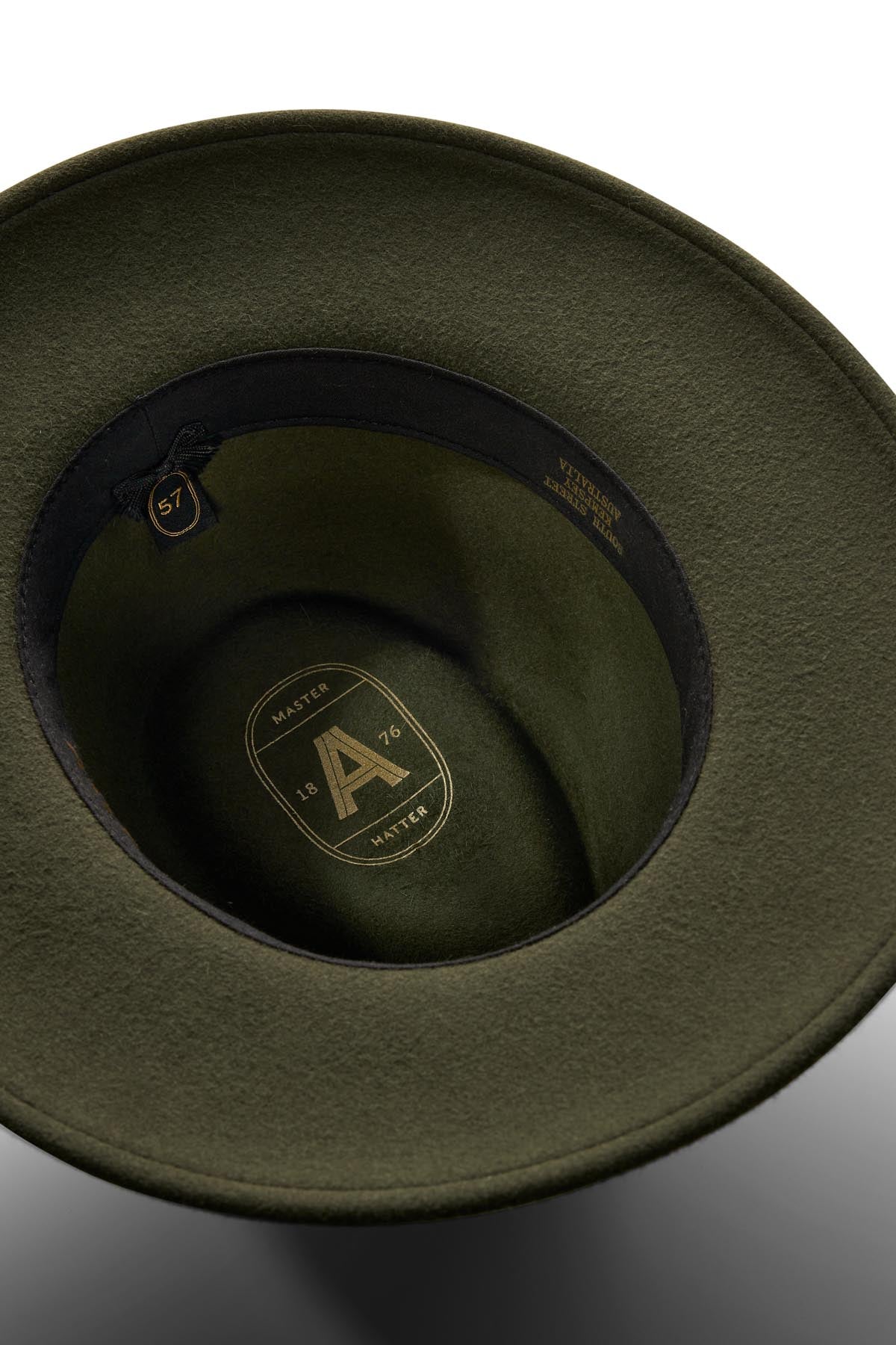 A close-up of the olive green Akubra Traveller hat's interior reveals its felt structure, black inner band, size label "57," and a gold logo with the letter "A," "MASTER," and "1876" on the lining.
