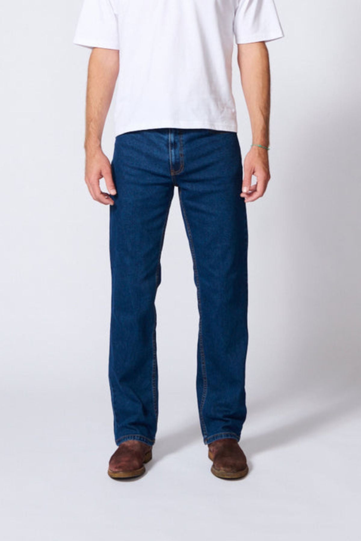 A person stands against a plain background, wearing a white T-shirt, AMCO Stretch Regular Fit Jean in indigo wash, and brown shoes. The photo is cropped above the shoulders, so their face is not visible.