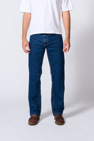 A person stands against a plain background, wearing a white T-shirt, AMCO Stretch Regular Fit Jean in indigo wash, and brown shoes. The photo is cropped above the shoulders, so their face is not visible.