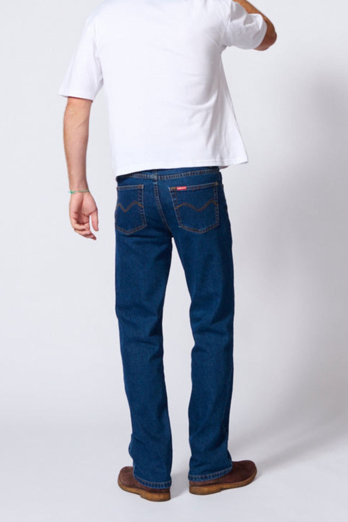 A person faces away from the camera, wearing a white t-shirt, brown shoes, and AMCO Stretch Regular Fit Jeans in indigo wash, against a plain light-colored background.