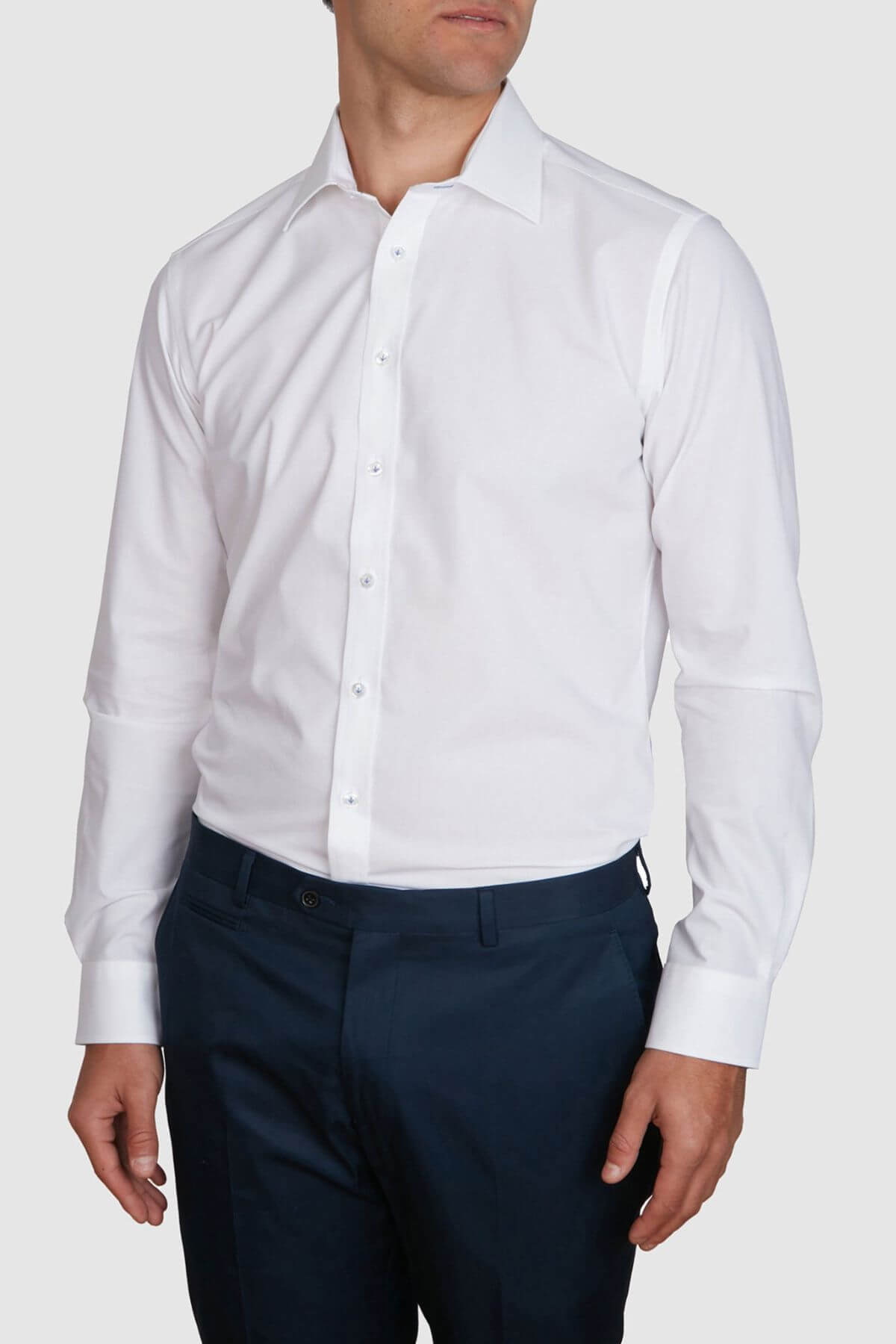 A man is wearing a white Abelard 4 Way Stretch Poplin Slim Fit dress shirt with the collar open and dark blue, slim fit trousers. The wrinkle-resistant shirt is long-sleeved and buttoned up, except for the top button. He is standing against a plain, light gray background.