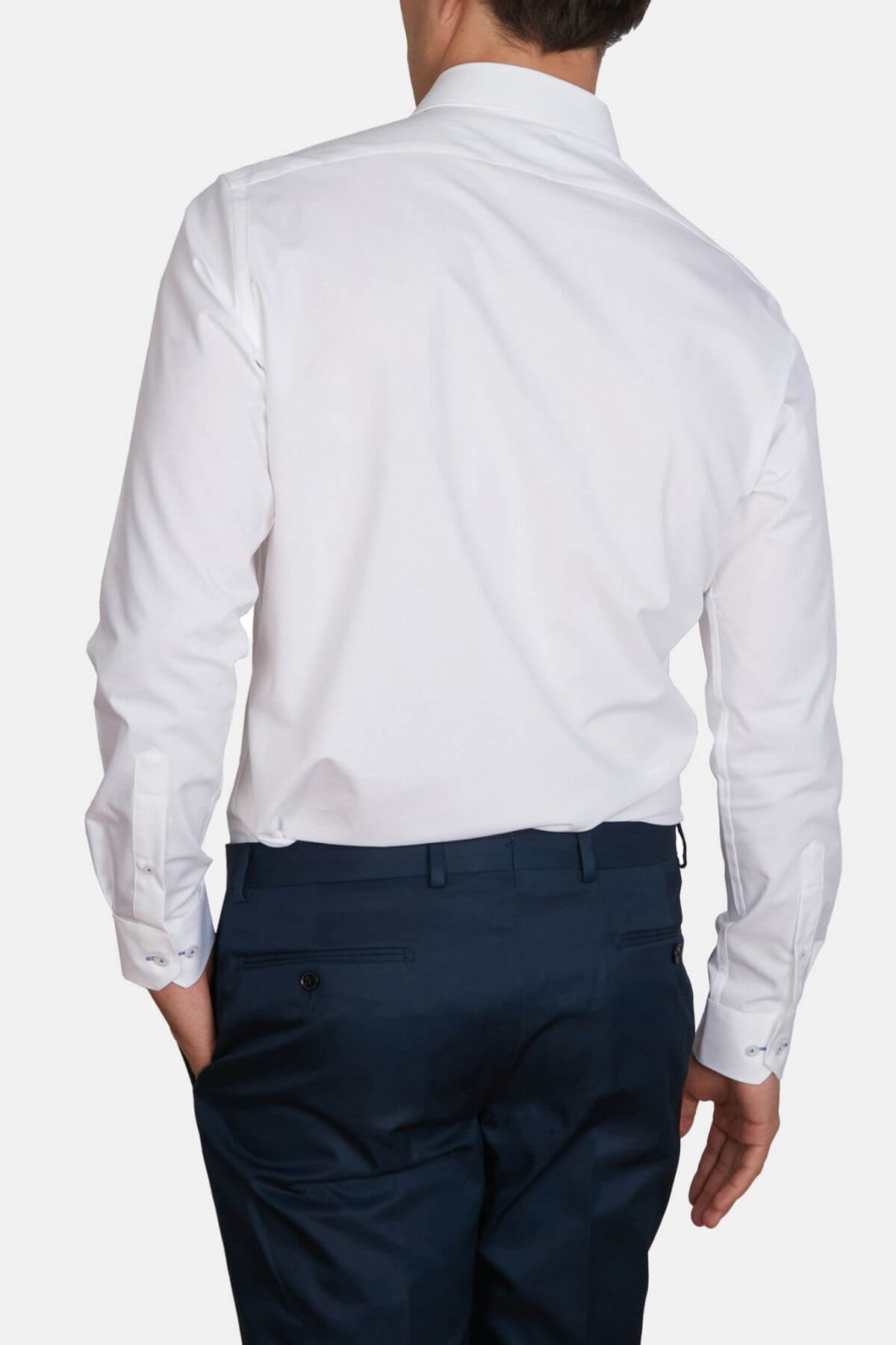 A person wearing a slim fit, white Abelard 4 Way Stretch Poplin Slim Fit dress shirt and dark blue trousers is shown from the back. The wrinkle-resistant shirt has long sleeves with cuffs, and the trousers have buttoned back pockets. The person stands with one hand in their pocket.