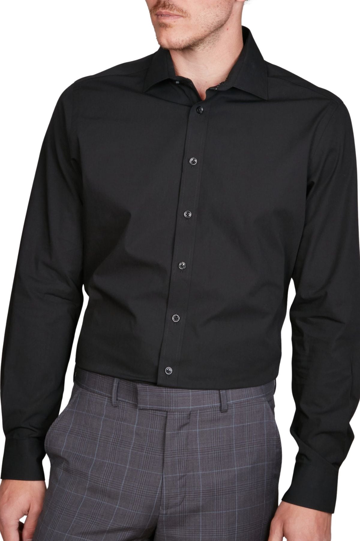 A man wearing the Abelard 4 Way Stretch Poplin Slim Fit black shirt and gray plaid dress pants is shown from mid-thigh up, facing slightly to the side with his face partially out of frame.
