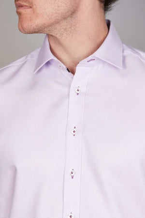 A close-up of a person wearing the Abelard Non Iron Cotton Twill Slim Fit Shirt. The light purple shirt is buttoned up, showcasing visible white buttons and buttonholes stitched in a slightly darker purple shade. Featuring a classic collar, this non-iron cotton shirt complements the individual's light complexion and short hair.
