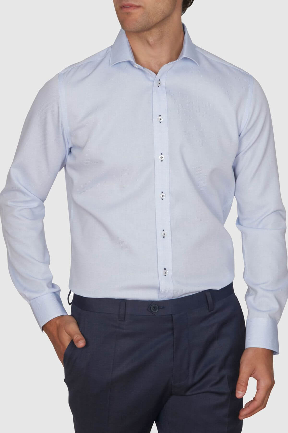 Someone wearing an Abelard Waffle Dobby Slim Fit Shirt in light blue, made of 100% cotton, paired with dark blue pants, standing against a plain background with one hand in a pocket. The shirt is neatly tucked in for a formal look.