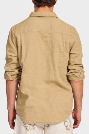 A man is seen from behind wearing the Academy Brand Harry Double Cloth Shirt, a light brown men’s casual shirt with rolled-up sleeves, paired with light floral shorts against a plain white background.