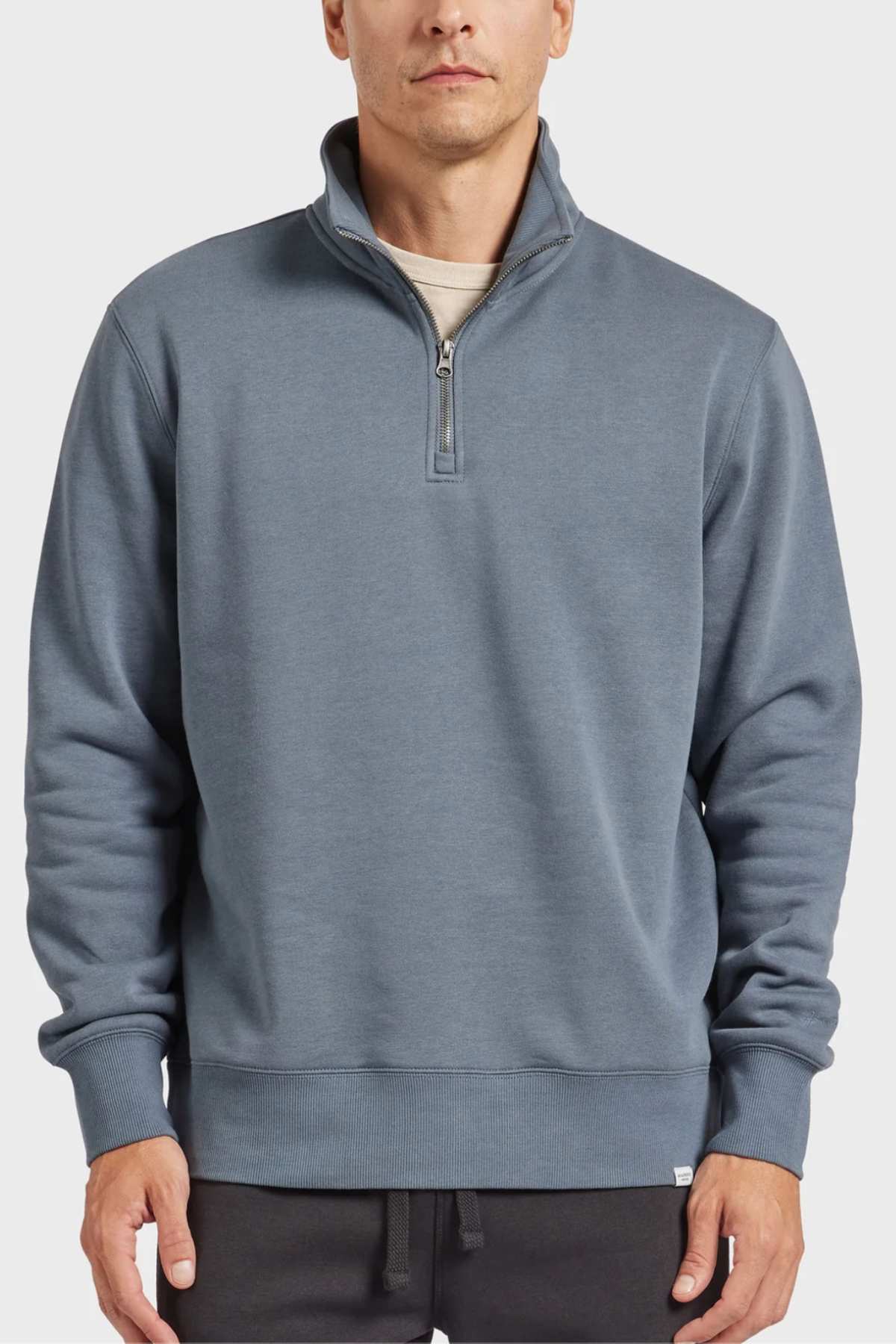 A man is wearing the Academy Brand 1/2 Zip Sweat in slate blue, which features tonal embroidery and a high collar. He pairs it with a light gray undershirt and dark pants, standing against a plain light gray background with his hands relaxed at his sides. The pullover boasts ribbed cuffs and hem, crafted from premium brushed fleece.